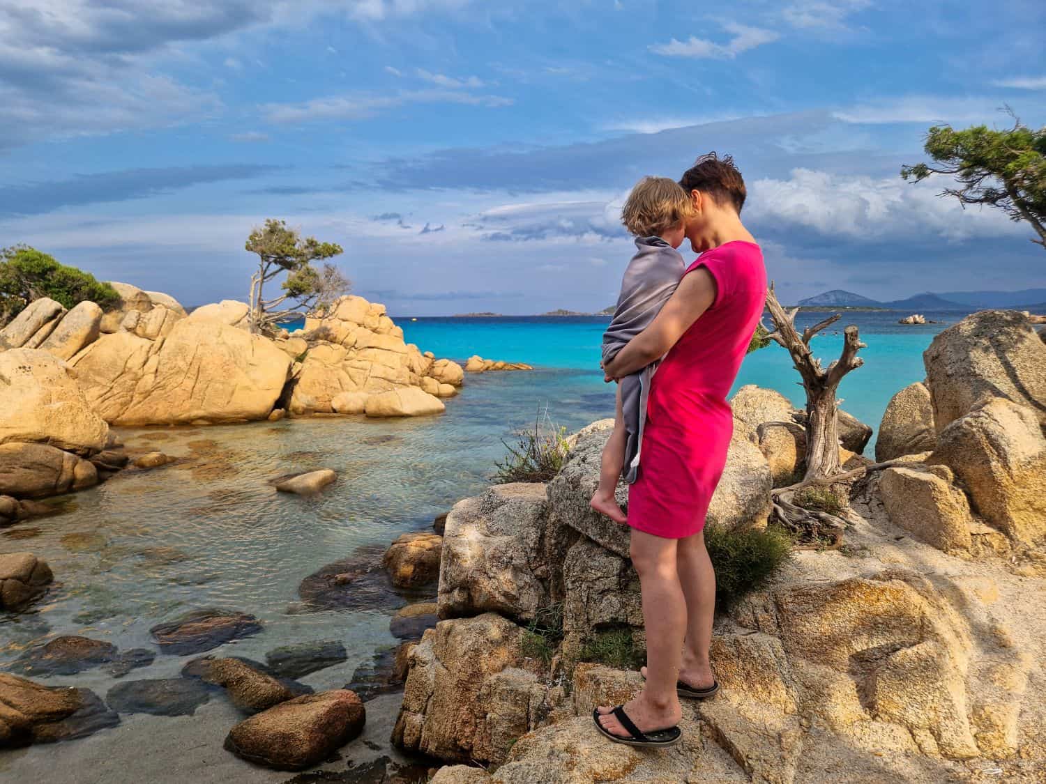 Emma and I on Cala Capriccioli beach in Sardinia Italy. there are huge boulders with sunlight on them and turquoise Mediterranean sea water in the background