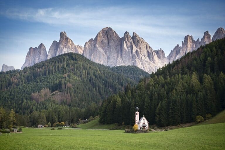 A small church with a bell tower stands in a lush green field, surrounded by dense pine forests. Behind the church, majestic jagged mountains rise against a clear blue sky, creating a serene and picturesque landscape.