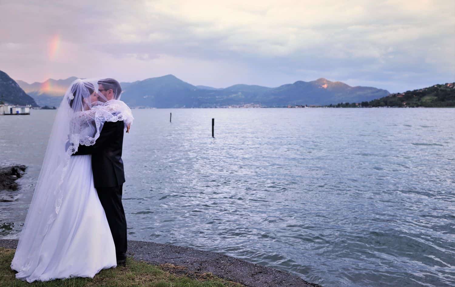 a bride and a groom - me and Michal - kissing next to lake Iseo with a rainbow behind us in the mountains