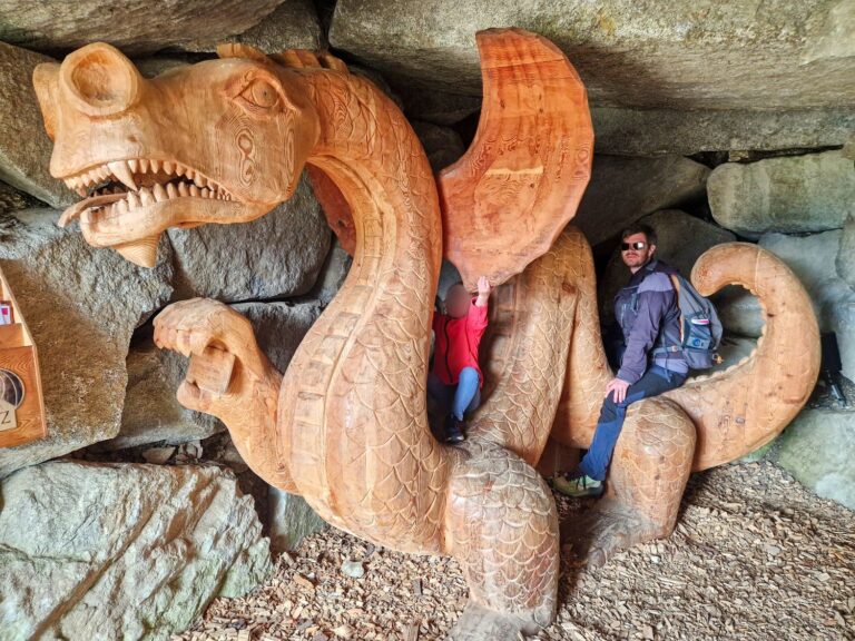 A man Michal and child Emma pose on a giant wooden dragon sculpture carved with detailed scales and sharp teeth inside a rocky cave shelter. The child sits beneath one wing while the man rests on the curled tail surrounded by large stone walls and wood chips on the ground.