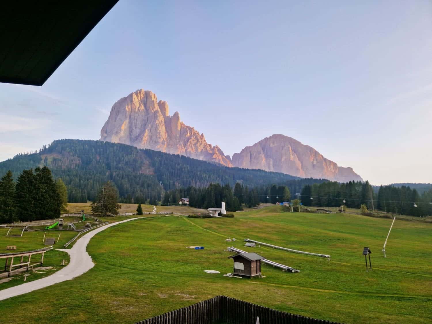 A scenic view of grassy fields leading to a majestic mountain range bathed in warm sunlight. The foreground shows meandering paths and small structures, with dense forests at the base of towering rocky peaks under a clear sky.