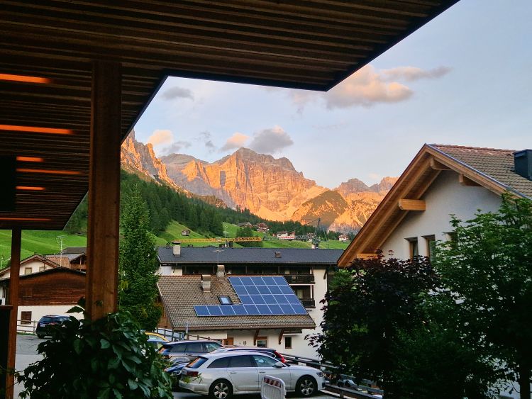 Things to Do in the Dolomites That Are Actually Worth It 40 Sunlit mountain peaks rise above a quiet alpine village San Cassiano in Alta Badia at sunset - enrosadira or the Alpenglow is in full force. Wooden balconies frame the view while rooftops with solar panels and parked cars sit below the glowing cliffs and scattered clouds. The view is from Hotel Diamant in San Cassiano.
