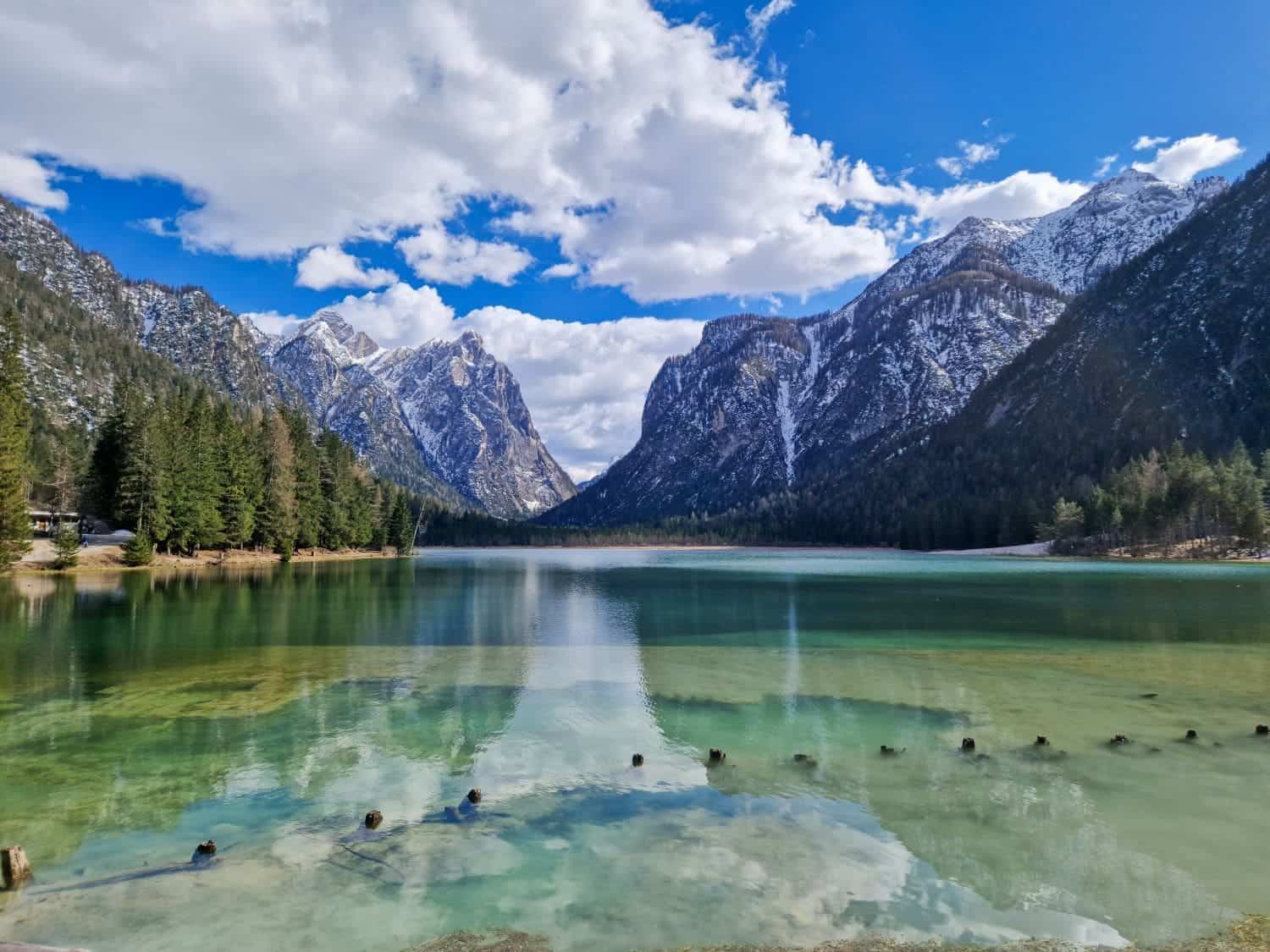 A tranquil lake surrounded by snow-capped mountains and evergreen trees under a partly cloudy sky. The water is clear, reflecting the blue sky and clouds, with logs partially submerged near the shore.