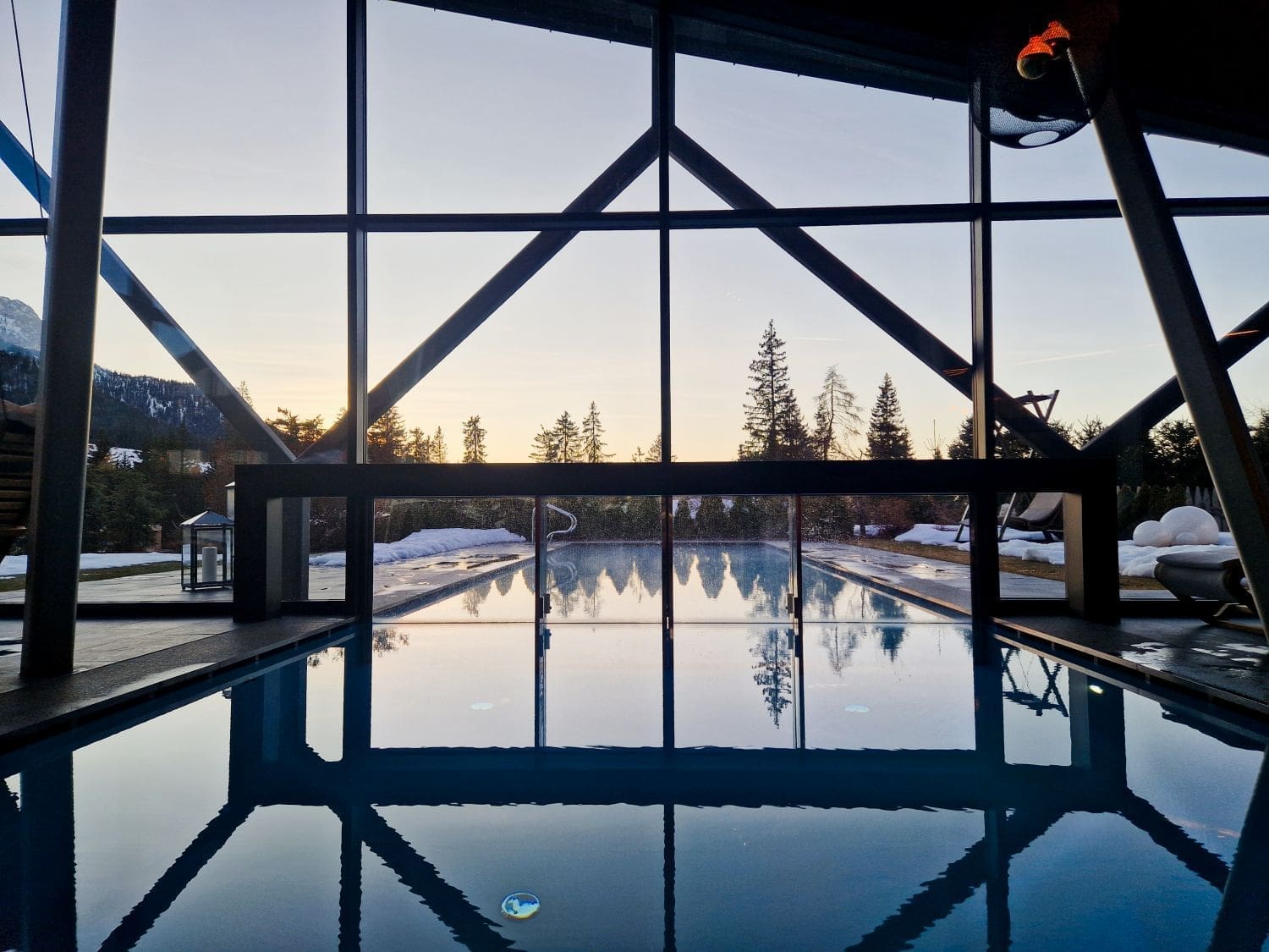 View from inside a modern indoor pool area with large windows showcasing an outdoor pool and scenic landscape. Snow-covered ground and trees are visible outside, with a sunrise or sunset illuminating the horizon and reflecting on the pool water.