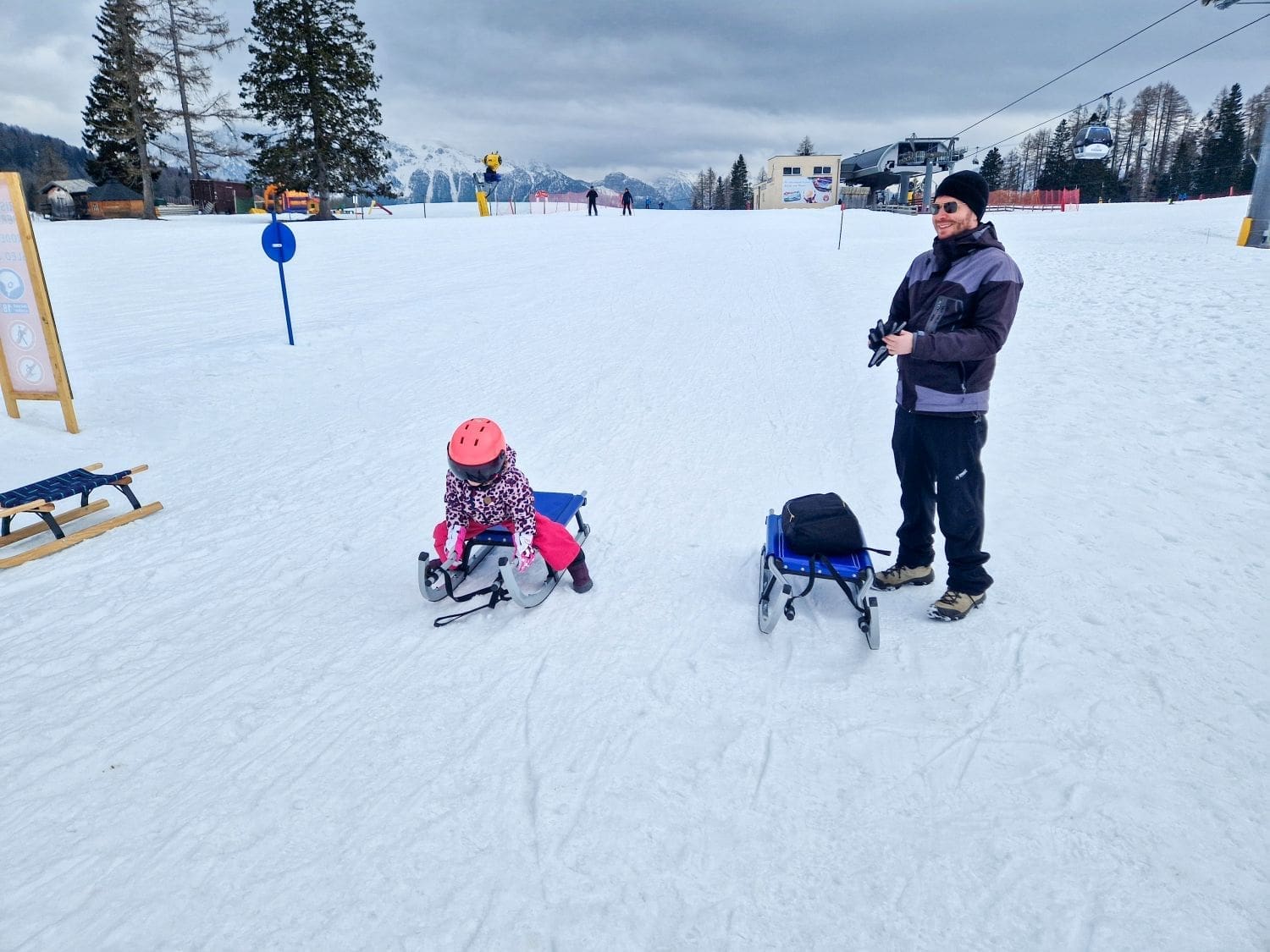 a kid sitting on a toboggan and a michal standing next to a toboggan before riding down the snowing mountain