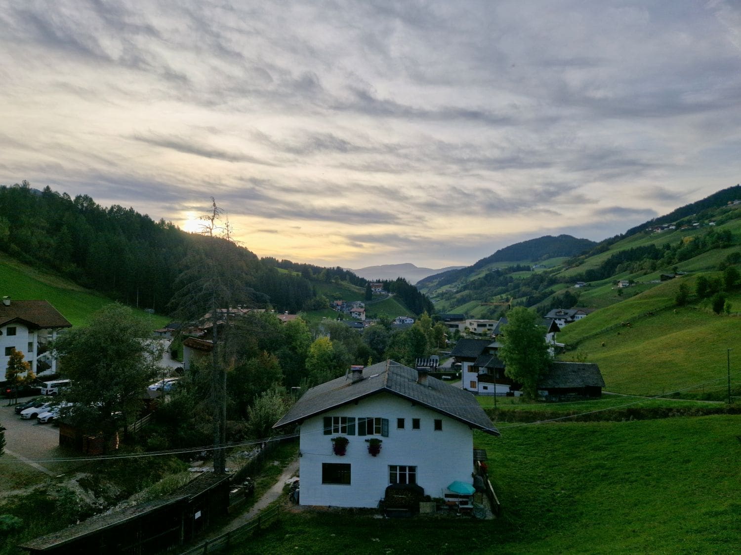 view of the val di funes valley during sunset. the hills are grassy, there's a white house in the foreground, and the sun is hiding behind the clouds