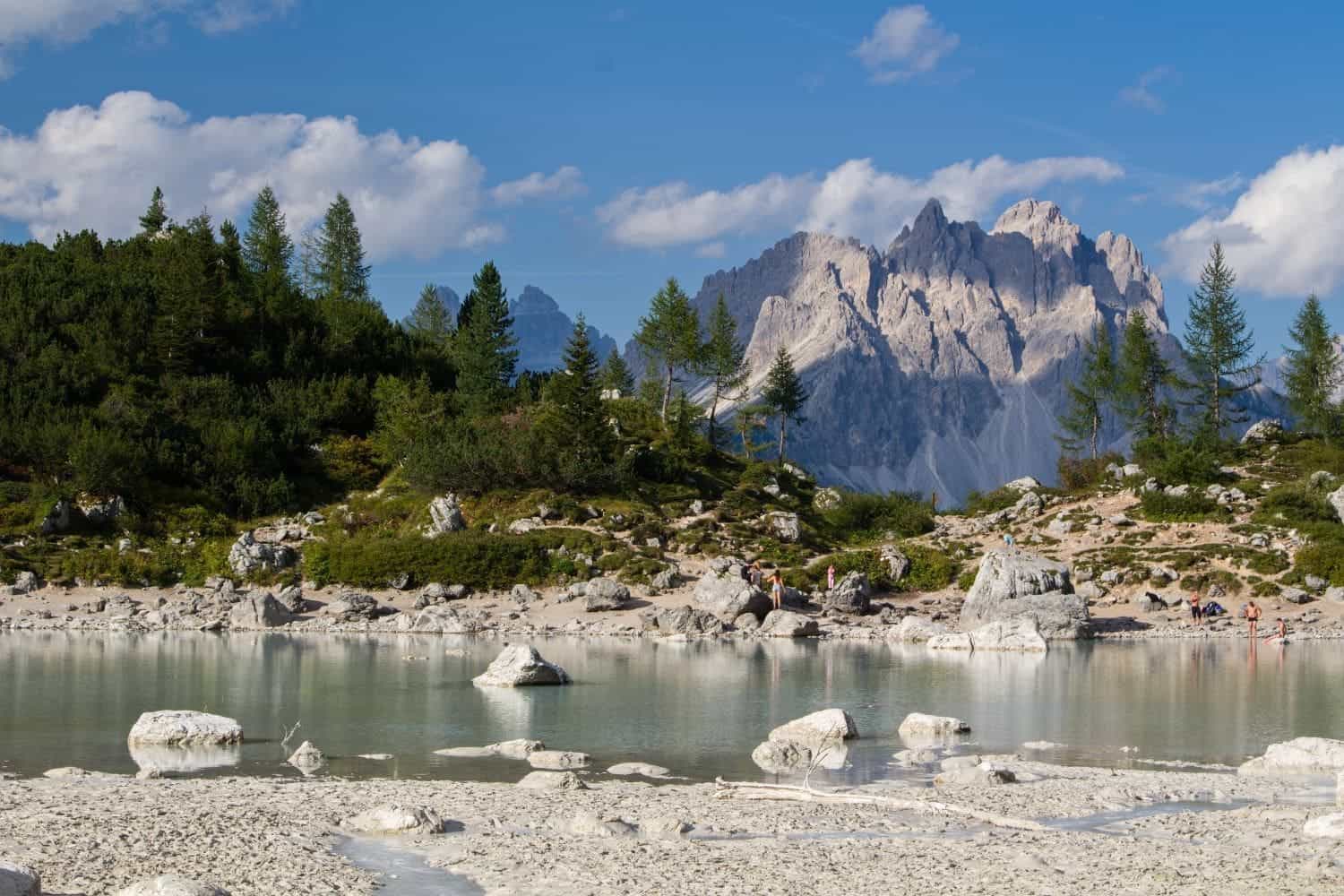 Lago di Sorapis Hike: Most Incredible Lake In The Dolomites 12 A tranquil alpine scene featuring a calm lake with large white rocks, surrounded by green trees. In the background, rugged mountains rise under a clear blue sky with scattered clouds.