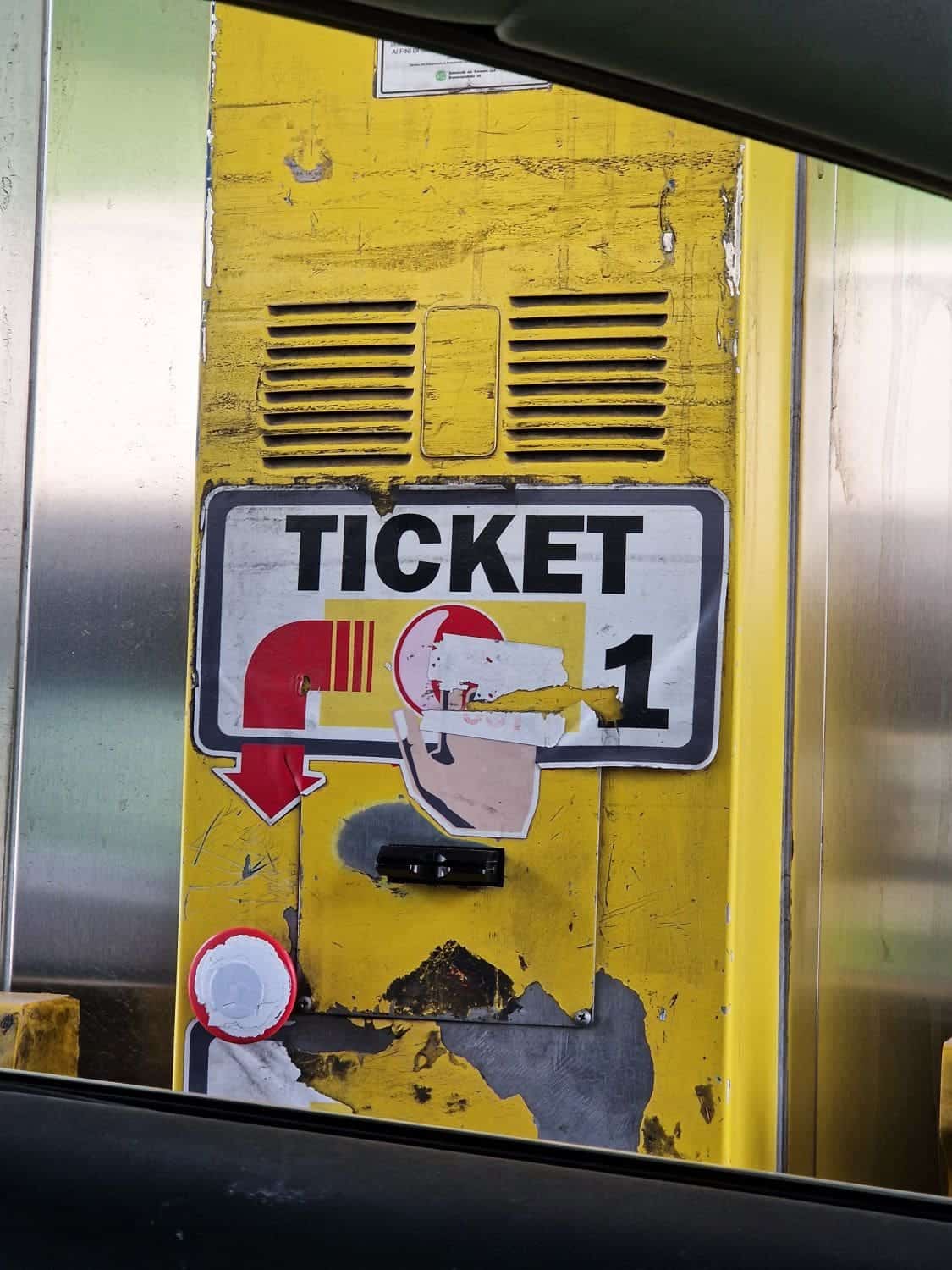 a yellow ticket machine on a highway in italy with a big writing "ticket" and a red arrow pointing to a red button