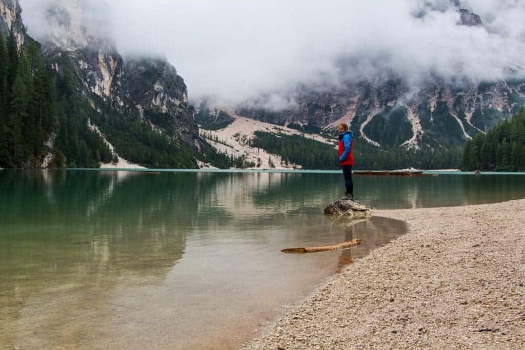 A person in a red and blue jacket stands on a rock by a tranquil lake surrounded by forested mountains. The sky is overcast with low-hanging clouds, creating a serene and misty atmosphere. The lake's surface reflects the mountains and trees.