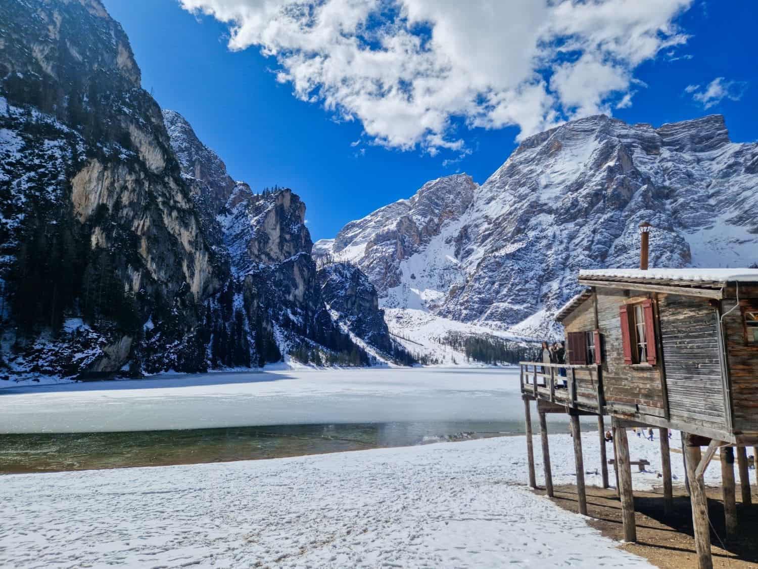 still mostly frozen lake braies in the dolomites in april, with a bit of greenish water visible. a boathouse on the right side of the image and snowy mountains in the background.
