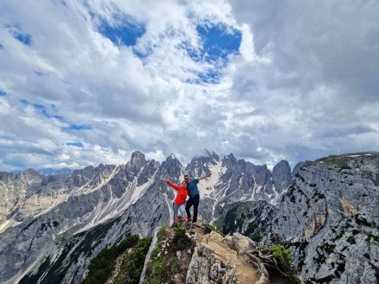 Two people stand on a rocky mountain peak with arms outstretched under a mostly cloudy sky. Jagged mountain peaks are visible in the background, and green shrubs dot the foreground.