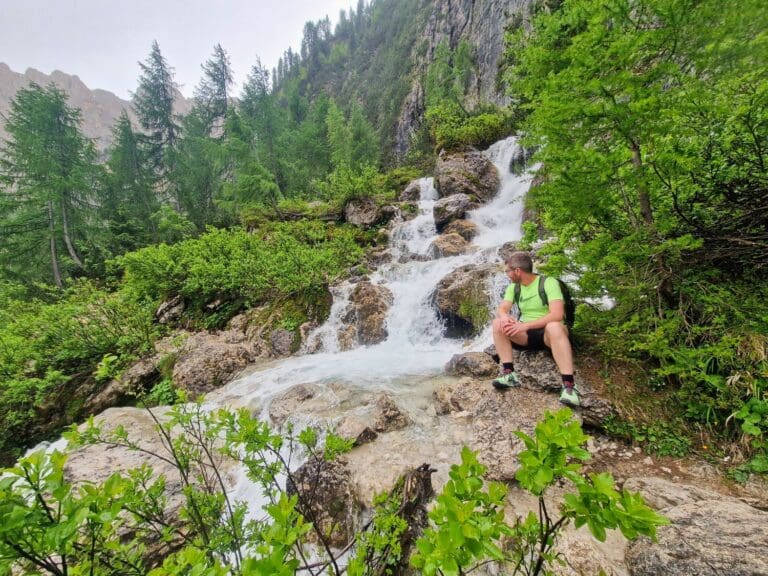 A person in a green shirt and shorts sits by a small waterfall in a lush, mountainous landscape. Surrounding them are green bushes and tall evergreen trees. The sky is overcast, and the scene is calm and serene.