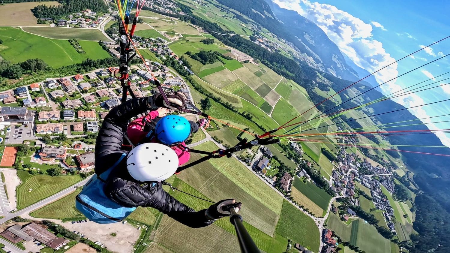 a view of tandem paragliding people in the Dolomites from Kronplatz with houses and fields on the ground and mountains in the background