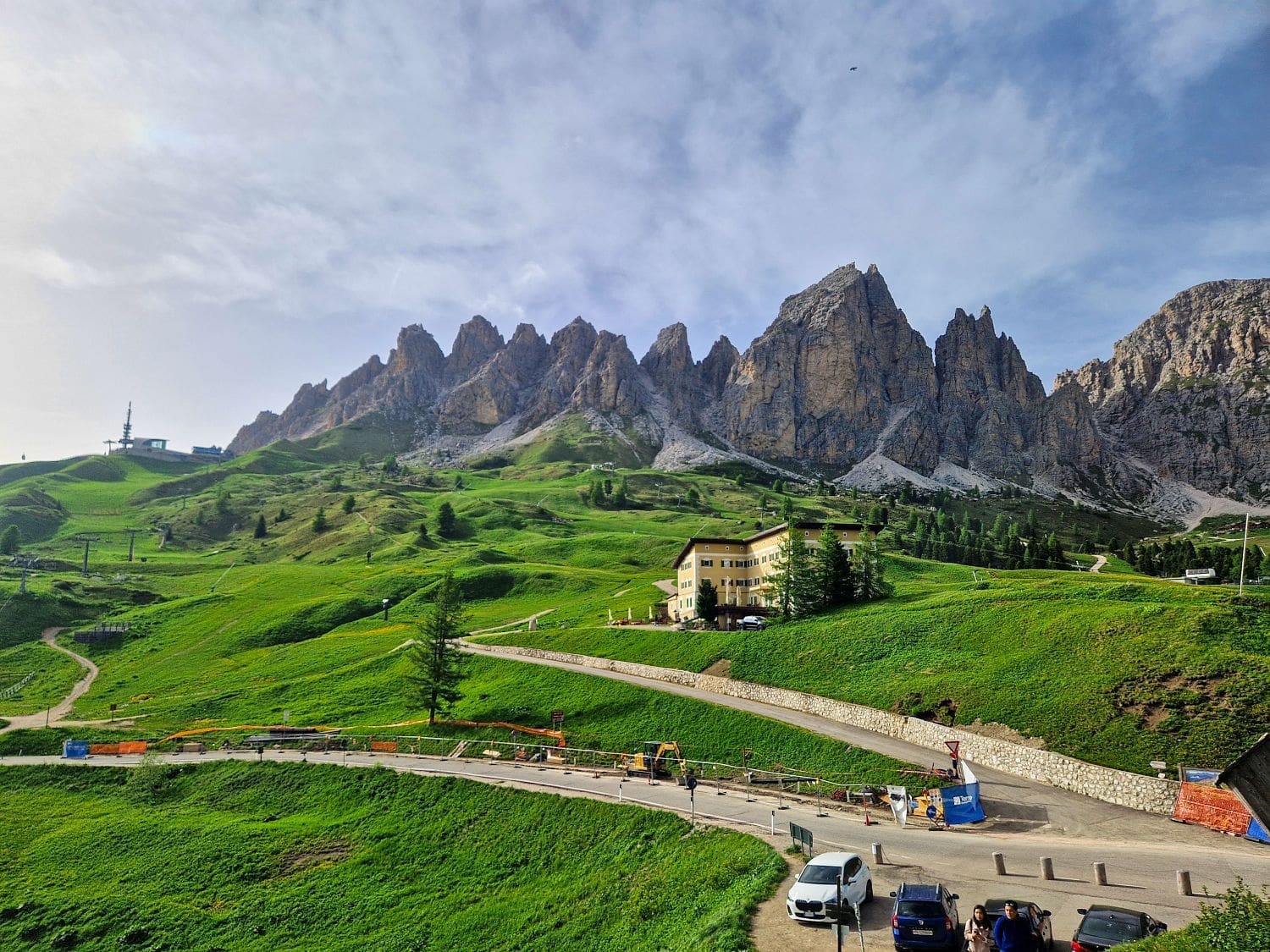 passo gardena in the Dolomites - mountains in the back, green meadows with a yellow hotel in the middle, and a parking lot in the foreground