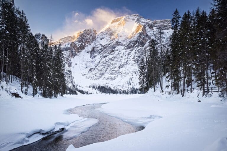 A snow-covered landscape with a winding river, surrounded by evergreen trees. In the background, tall mountains are illuminated by golden sunlight at their peaks under a clear blue sky.