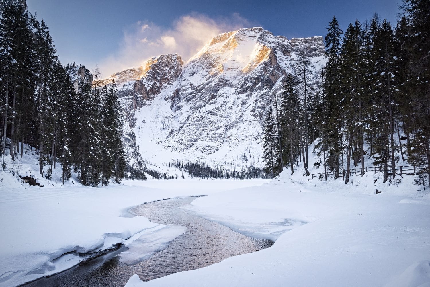 lago di braies at sunrise. the sun is shining on the snowy mountains, and there's a river with snow banks on the sides.