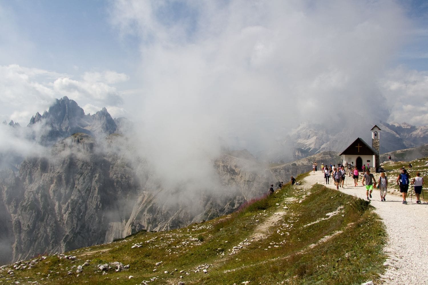 tre cime di lavaredo in august in the dolomites with a little church, cloudy mountains, and lots of people on the trail