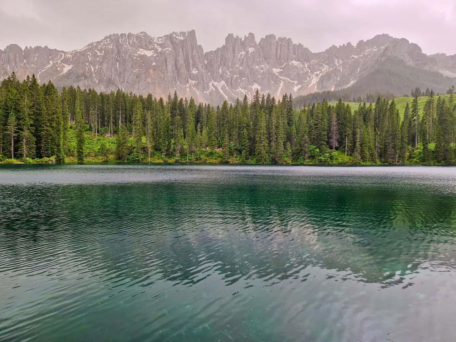 A serene lake carezza with calm, reflective waters is surrounded by dense, tall pine trees. In the background, jagged gray mountains partially covered with patches of snow rise into a cloudy sky, creating a peaceful and picturesque landscape.