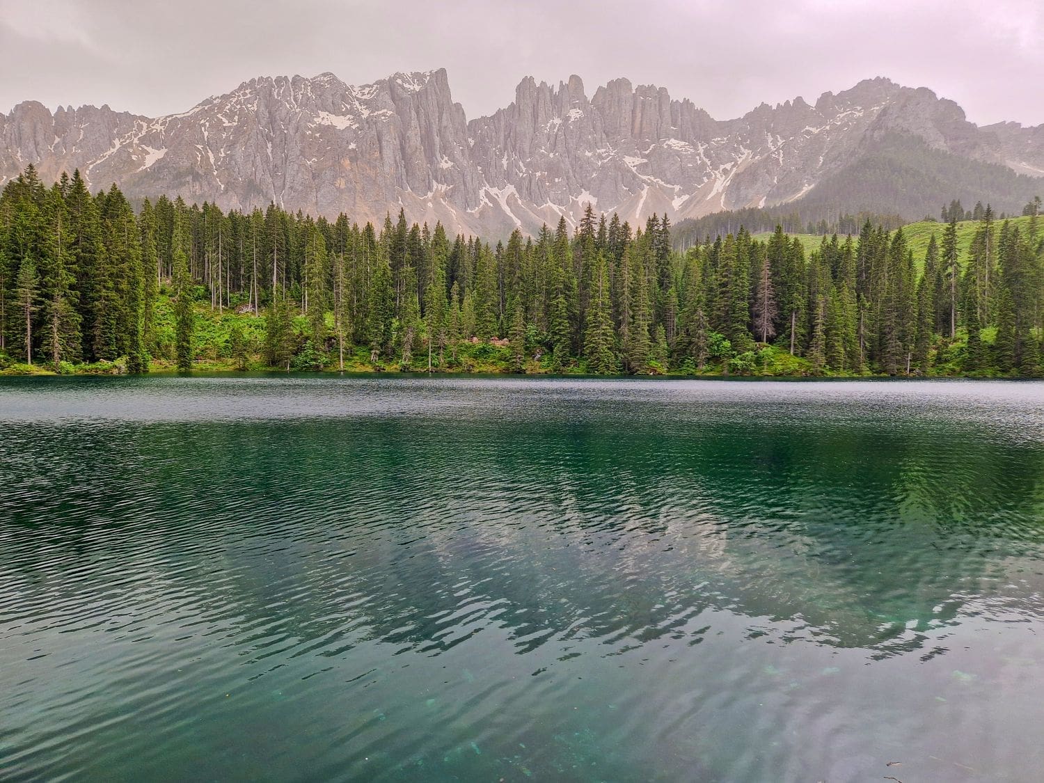 A serene lake carezza with calm, reflective waters is surrounded by dense, tall pine trees. In the background, jagged gray mountains partially covered with patches of snow rise into a cloudy sky, creating a peaceful and picturesque landscape.