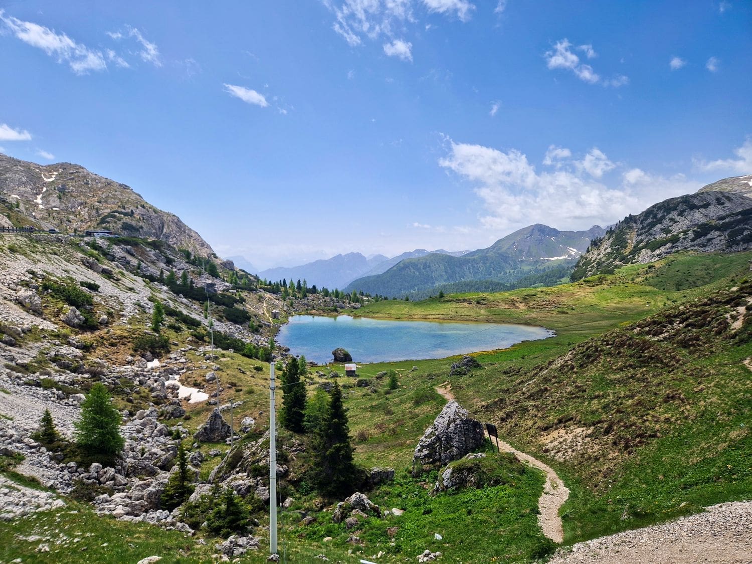 A scenic view of a mountainous landscape featuring a clear blue lake Valparola surrounded by green meadows and rocky terrain. A pathway leads to the water, with scattered trees and distant, snow-capped peaks under a partly cloudy sky.