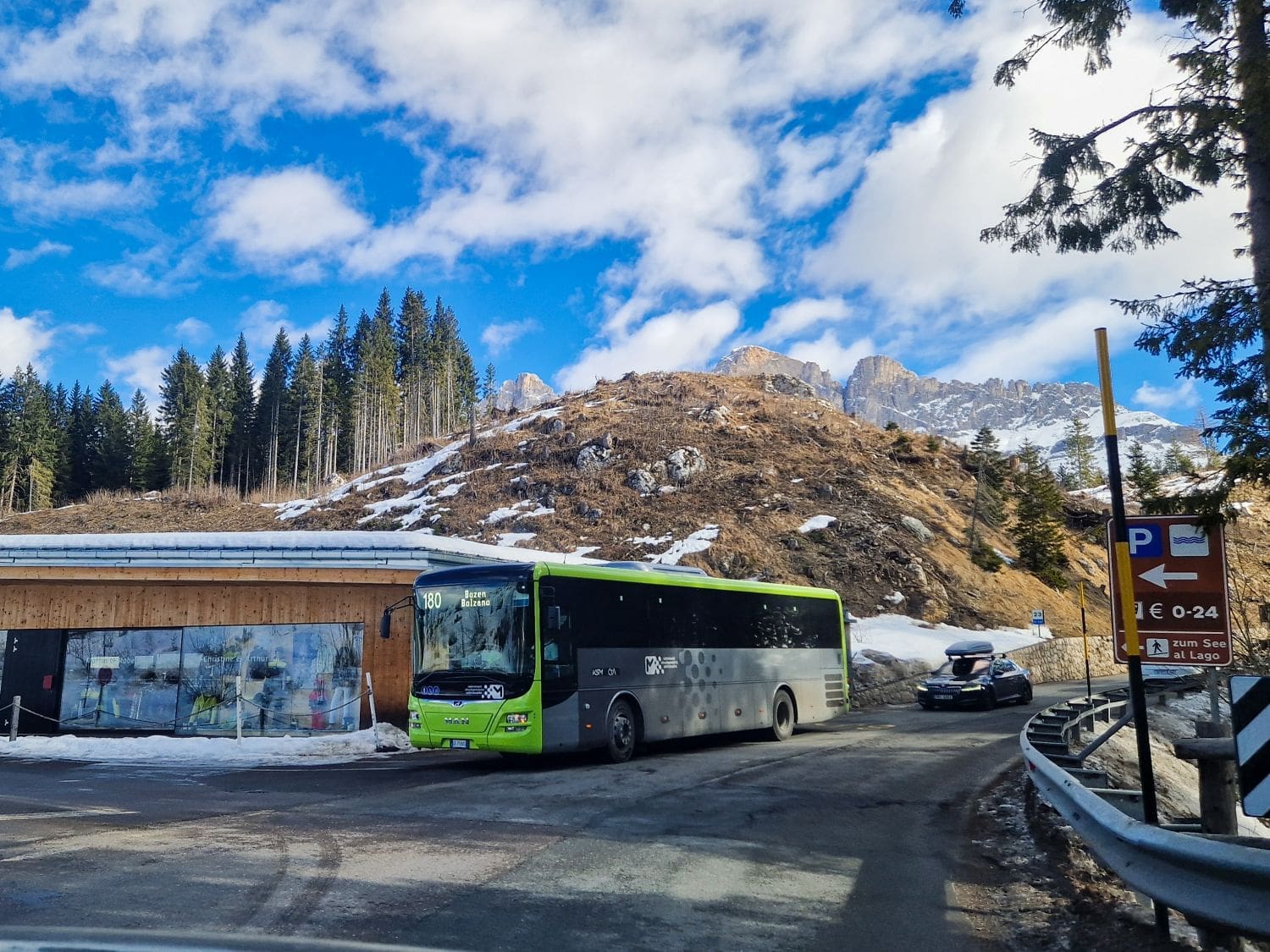 A green and gray bus is on a mountain road with snow and pine trees around. A small building with a mural is on the left. Mountains and a partly cloudy blue sky are in the background. Road signs indicate directions and parking information.