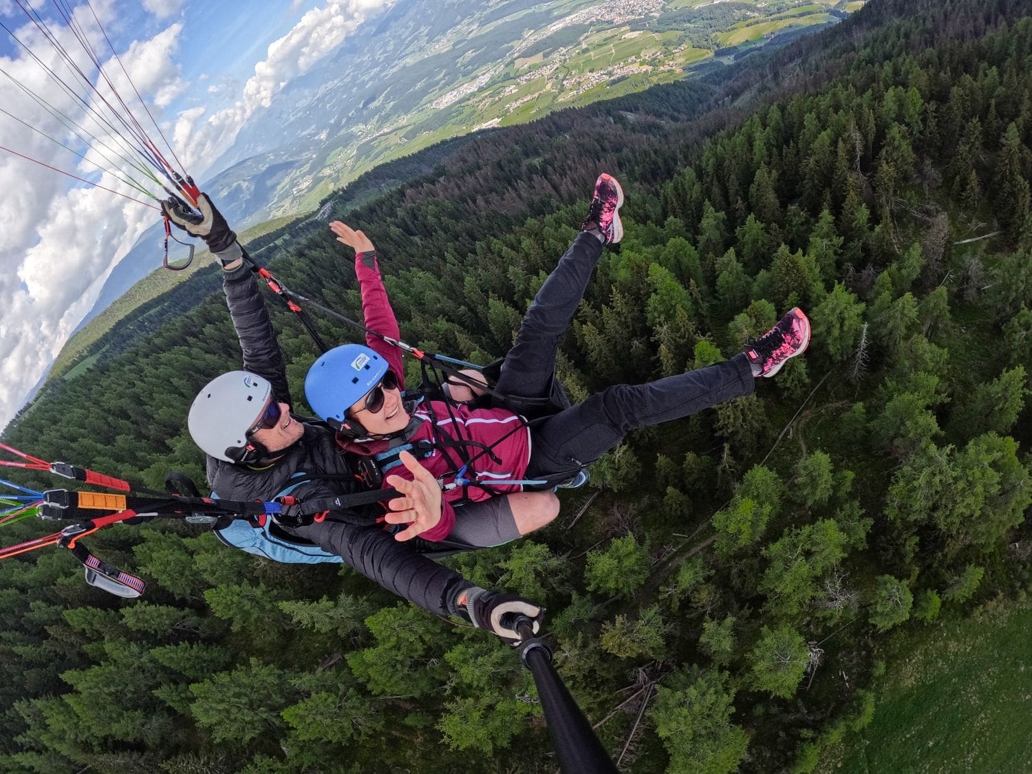 Two people are paragliding in the Dolomites over a lush forest, with one person wearing a blue helmet and the other a red helmet. They are tandem paragliding, with the instructor in the rear. Both have their arms outstretched, and a scenic landscape of trees and hills is visible below.