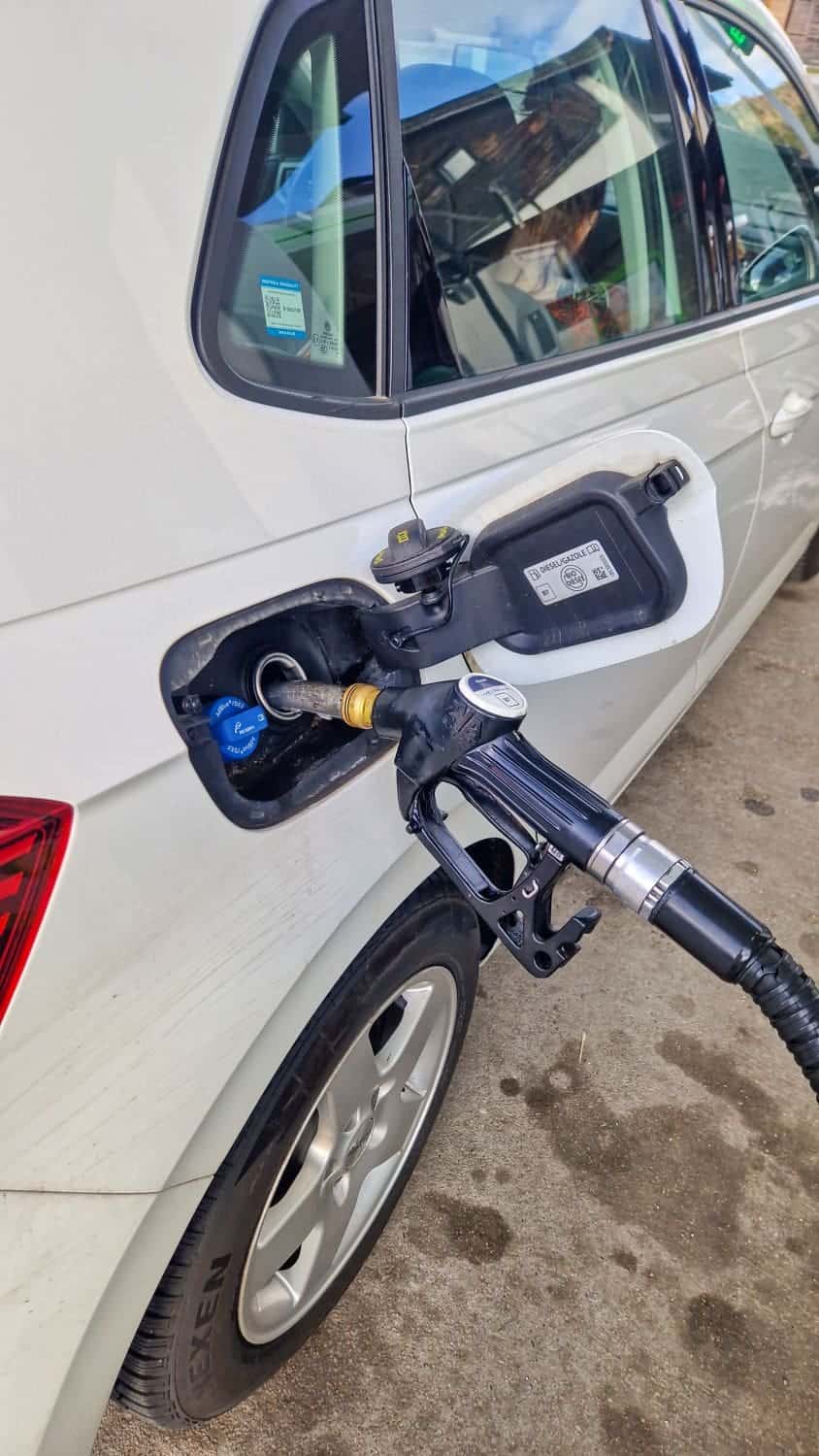 A white car is refueling at a gas station. The fuel pump nozzle is inserted into the car's gas tank, and the car door is open. The scene is captured in daylight.