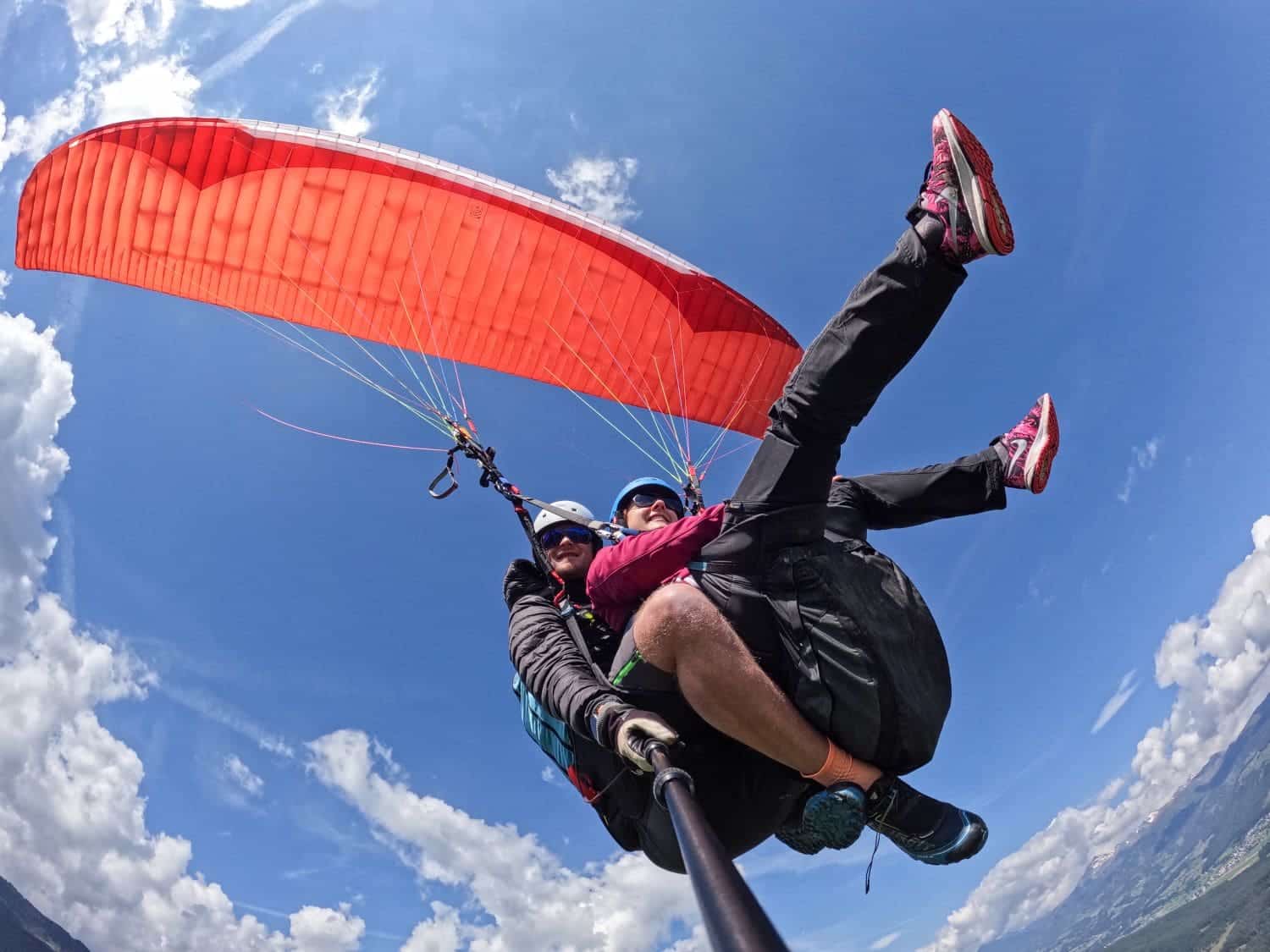 Two people are tandem paragliding high in the sky with a red paraglider. They are wearing helmets, harnesses, and sunglasses, soaring above a landscape of green hills under a blue sky scattered with white clouds. Paragliding in the Dolomites, they appear to be enjoying the adventure immensely.