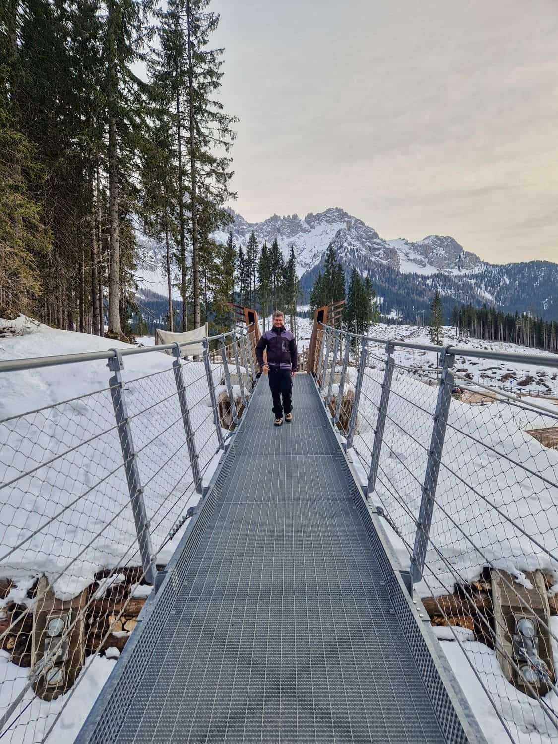 A person walks across a metal suspension bridge surrounded by snow-covered terrain and pine trees near Lake Carezza. Mountains with a light dusting of snow are visible in the background under an overcast sky.