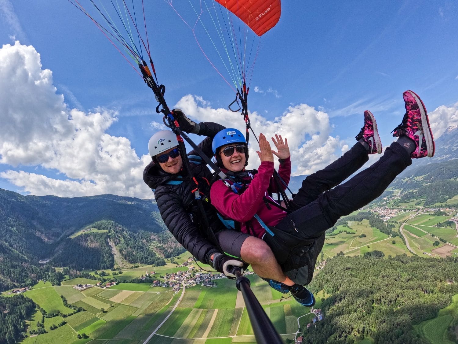 Two people are tandem paragliding on a sunny day over the scenic landscape of green fields, hills, and a small village in the Dolomites. They are wearing helmets and harnesses, and both appear to be enjoying the experience, with one holding a selfie stick to capture the moment.