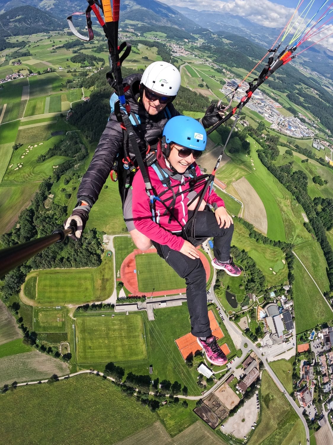 Two individuals are tandem paragliding high above a picturesque landscape of green fields, roads, and buildings in the Dolomites. Both are wearing helmets and harnesses; the back person is holding a selfie stick to capture the moment. The sky is blue with scattered clouds, and mountains are visible in the distance.