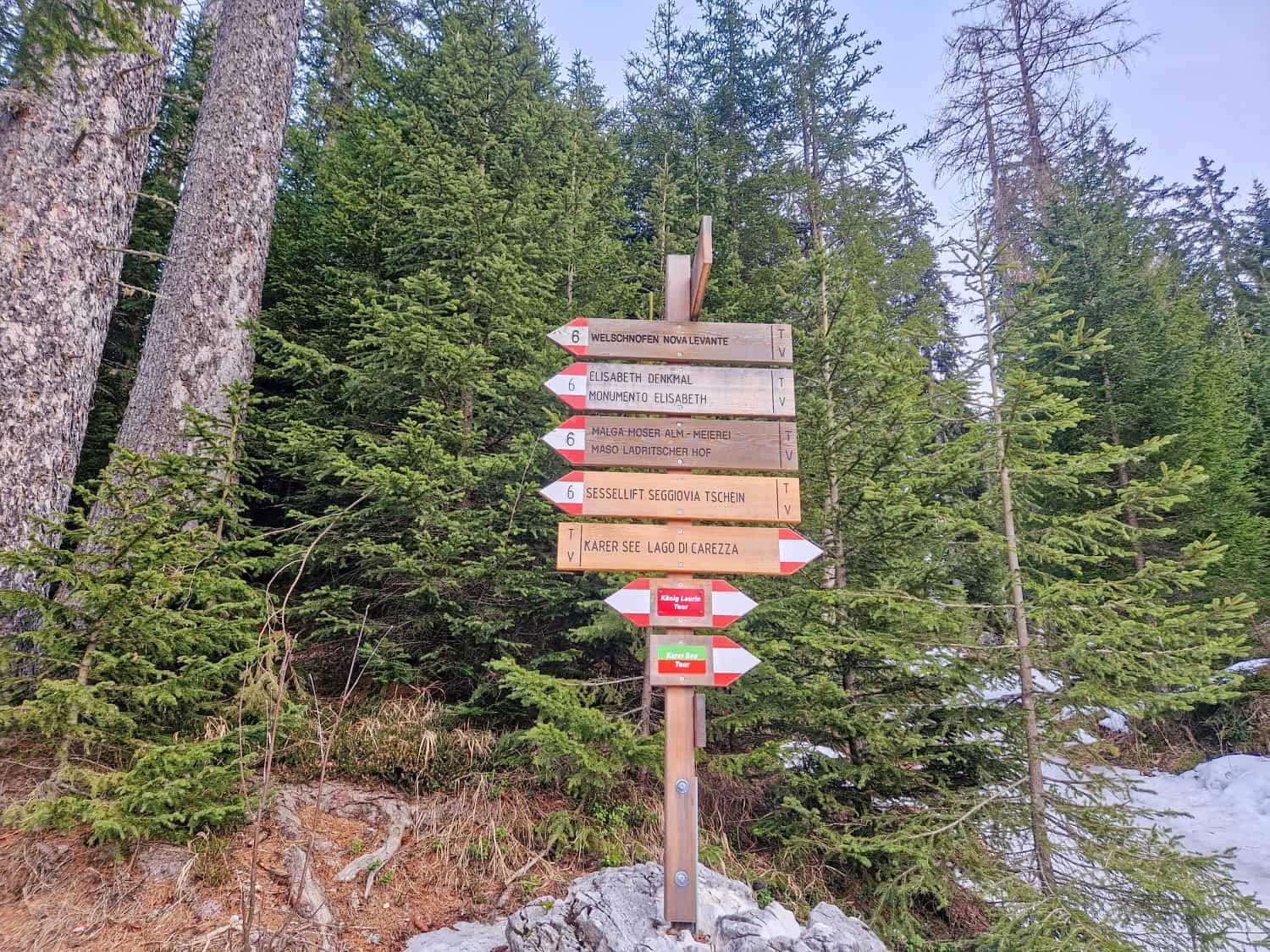 Wooden signpost surrounded by trees, showing various hiking trail directions and landmarks, including Malga Costa, Elsenora Tower, Sentiero Joch Trail, and Lago di Carezza. Snow patches are visible on the ground around the sign.