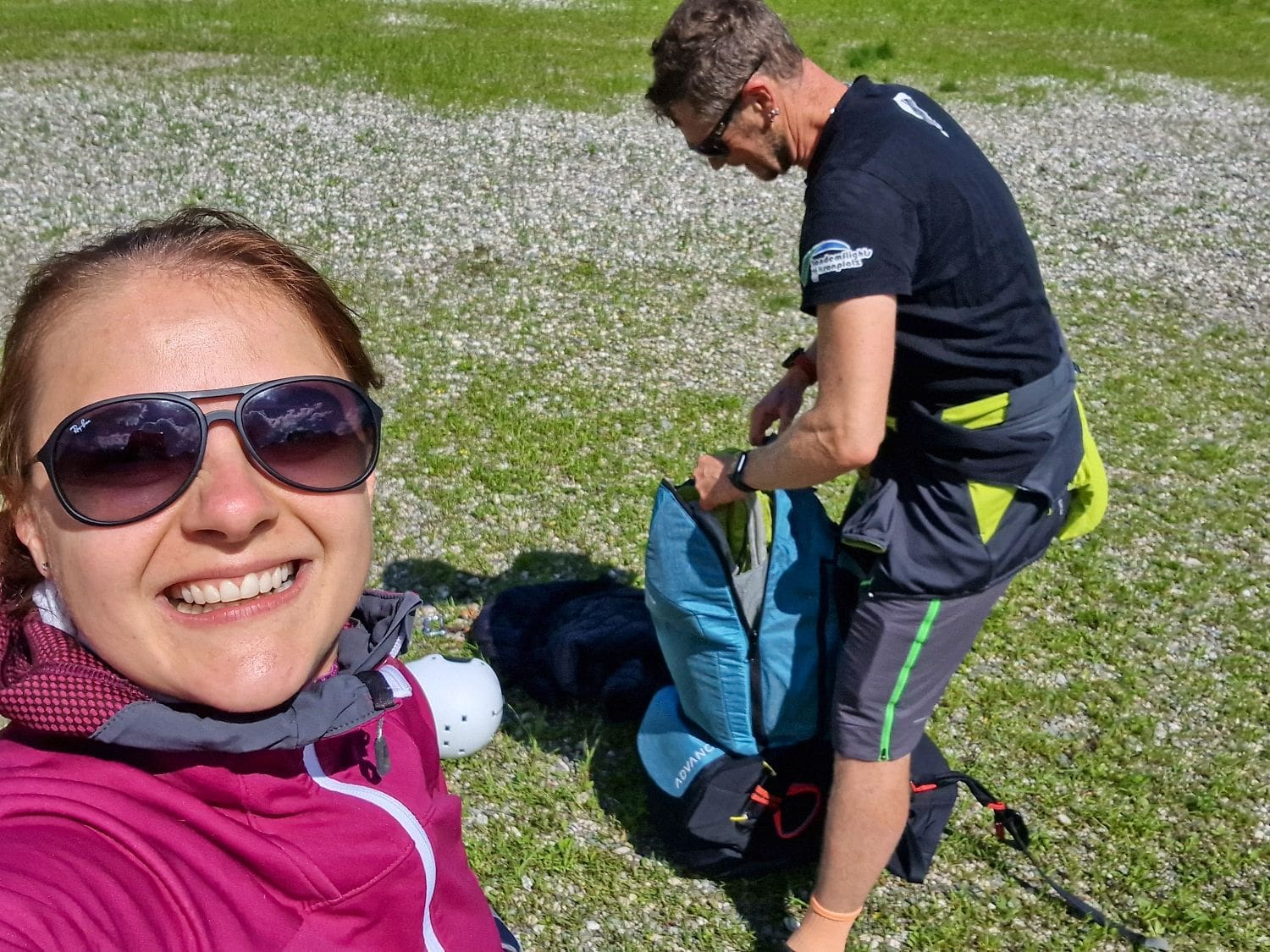 A woman in sunglasses smiles at the camera, wearing a purple jacket. Next to her, a man in a black shirt and shorts is bent over, packing a blue and black bag. They are outdoors on a grassy area with patches of small white flowers, preparing for tandem paragliding in the Dolomites under sunny weather.