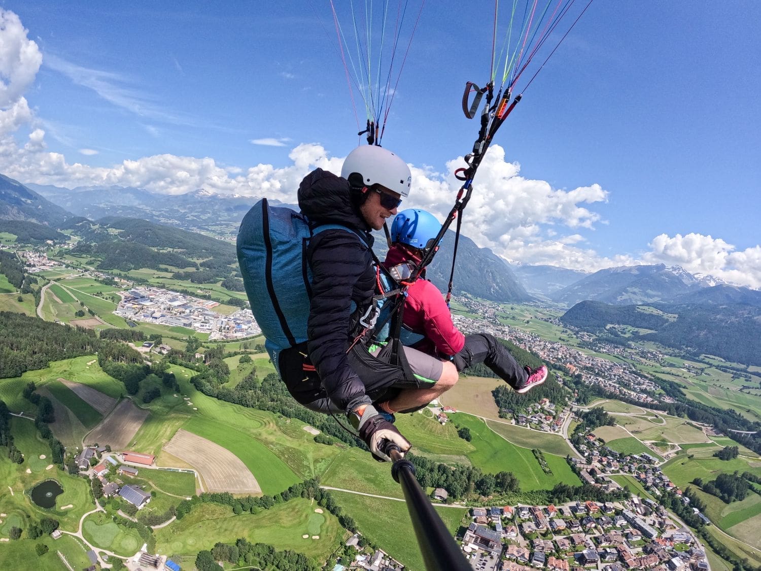 Two people are tandem paragliding in the Dolomites over a scenic landscape featuring green fields, a small town, and mountains in the distance. The individuals wear helmets and safety gear, with colorful parachute lines extending above them. One person is holding a selfie stick to capture the moment.