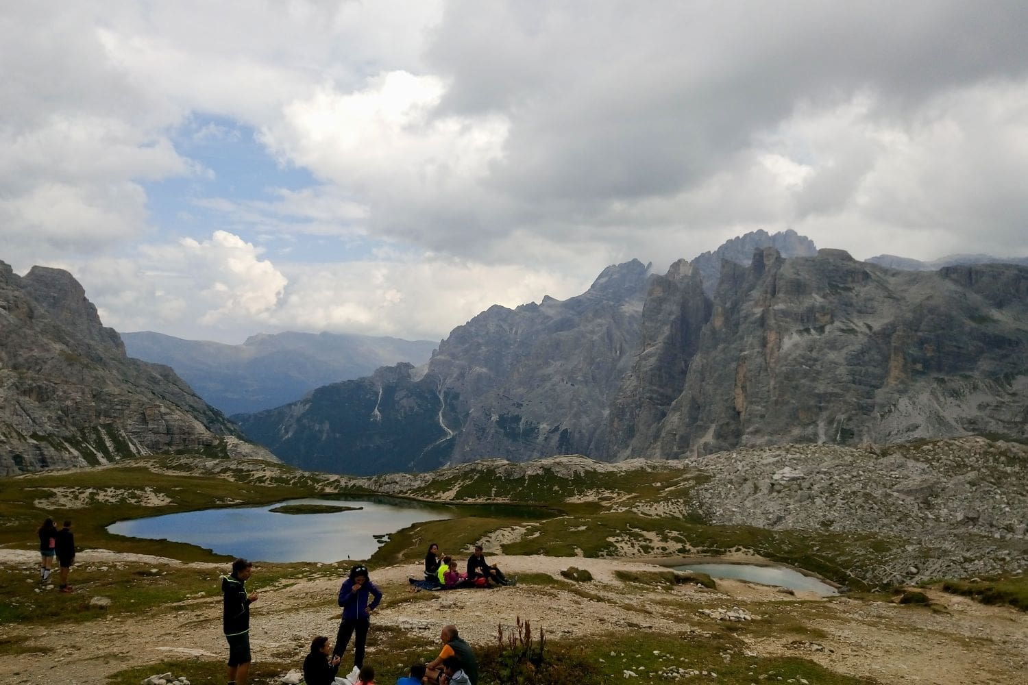 A cloudy day in a mountainous region with rugged peaks and valleys. In the foreground, several people are gathered near a grassy area with scattered rocks. Two blue alpine lakes are visible, and the background features a dramatic landscape of steep cliffs and ridges.