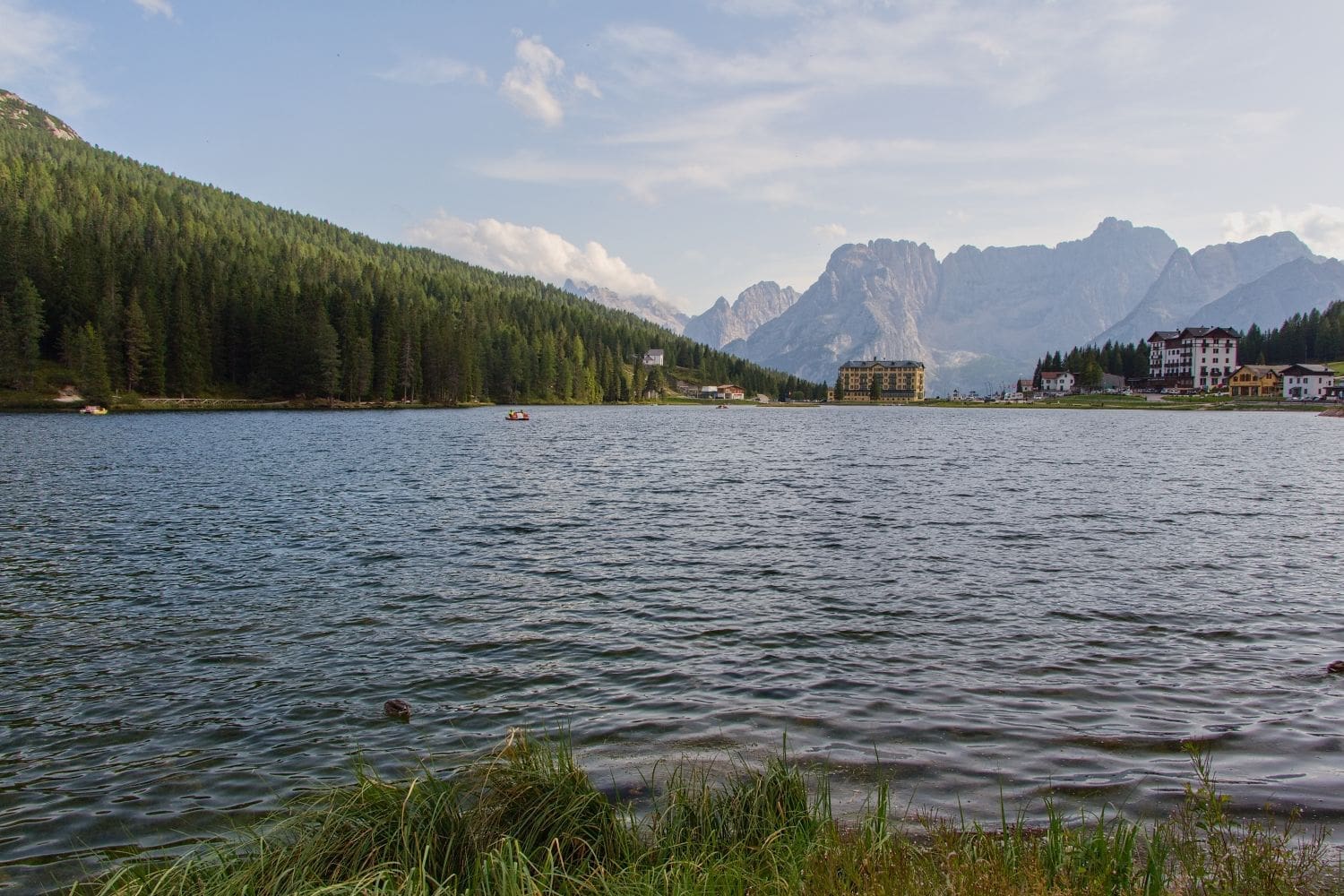 A serene lake bordered by dense forests and overlooked by towering mountains. In the distance, a few buildings, including a prominent large yellow structure, sit near the water’s edge. Small boats are visible on the calm water, enhancing the tranquil ambiance.