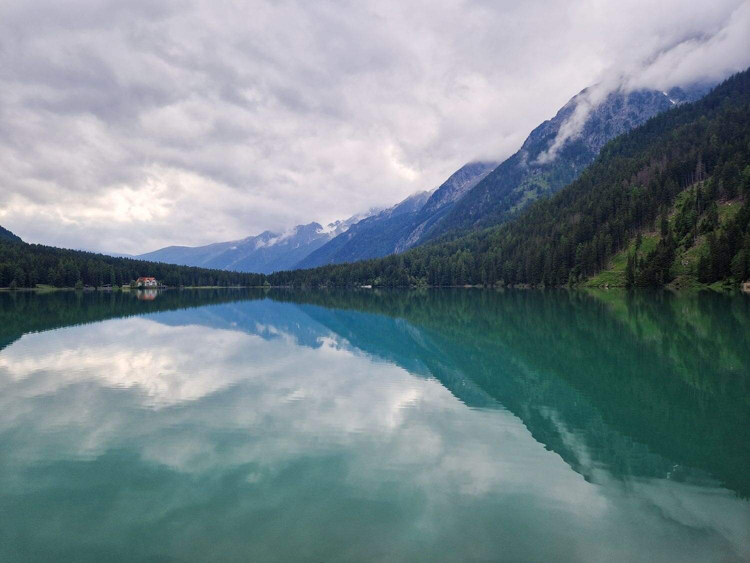 A serene mountain lake surrounded by lush green forests. The calm, turquoise water reflects the cloudy sky and the towering mountains in the background, which are partially covered in mist. A hotel sits on the distant shore to the left.