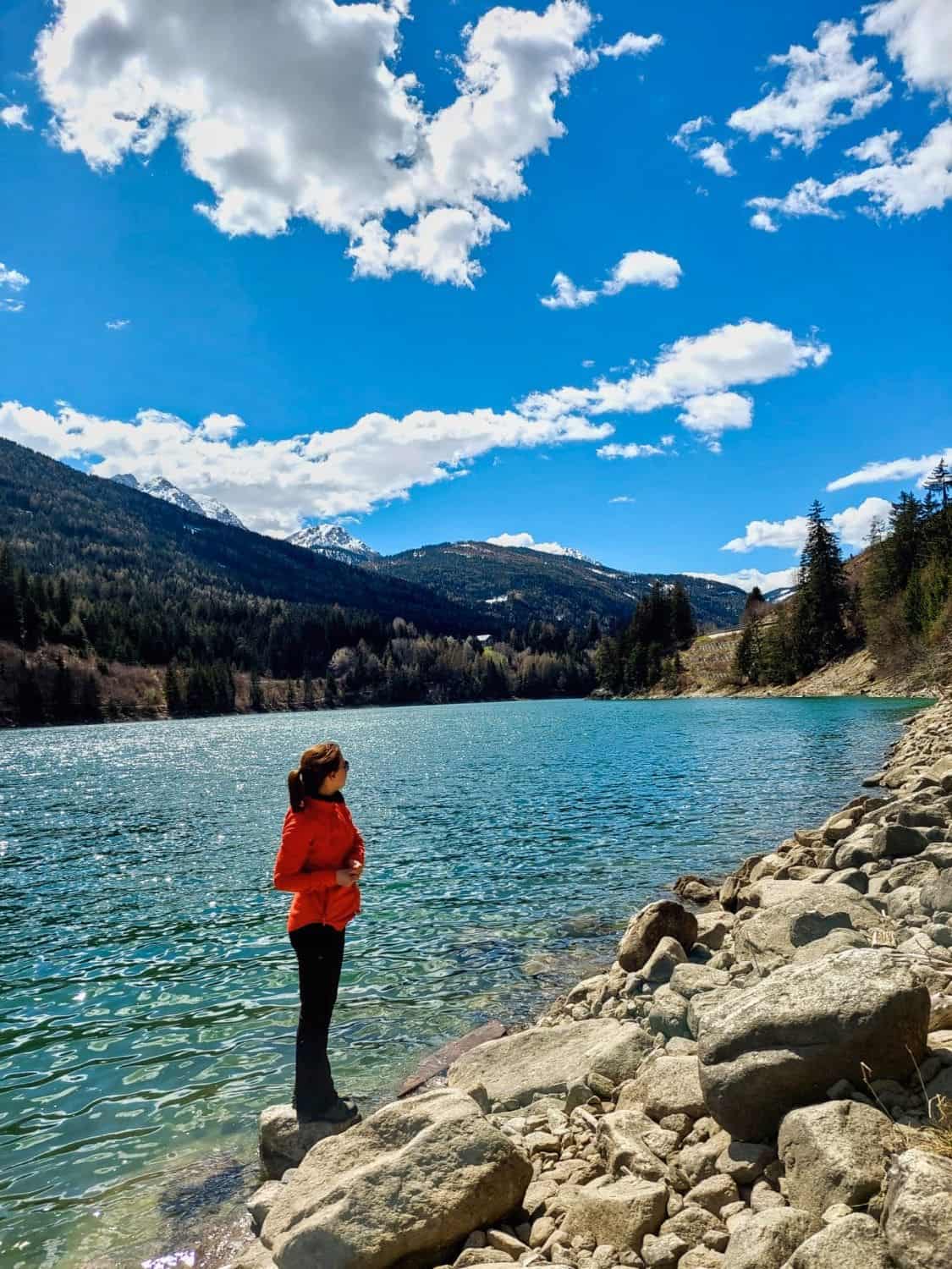 A person in a red jacket stands on rocks by a turquoise lake surrounded by forested mountains under a bright blue sky with scattered clouds.