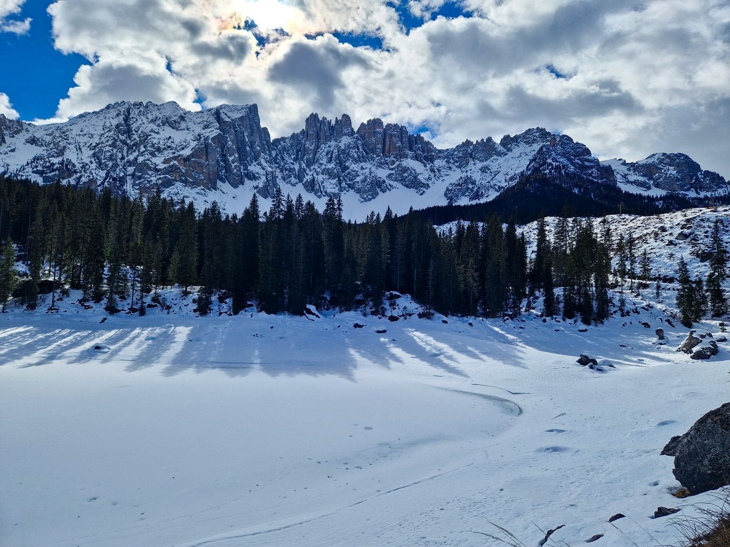 A snowy landscape featuring a frozen Lago di Carezza, surrounded by snow-covered rocks and pine trees. In the background, a rugged, snow-dusted mountain range is partially illuminated by sunlight breaking through scattered clouds in a blue sky.