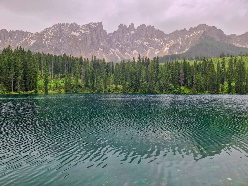 A serene mountain lake with clear blue-green water is surrounded by green pine trees. In the background, rugged, snow-capped mountains rise under a cloudy sky, creating a tranquil and picturesque scene.