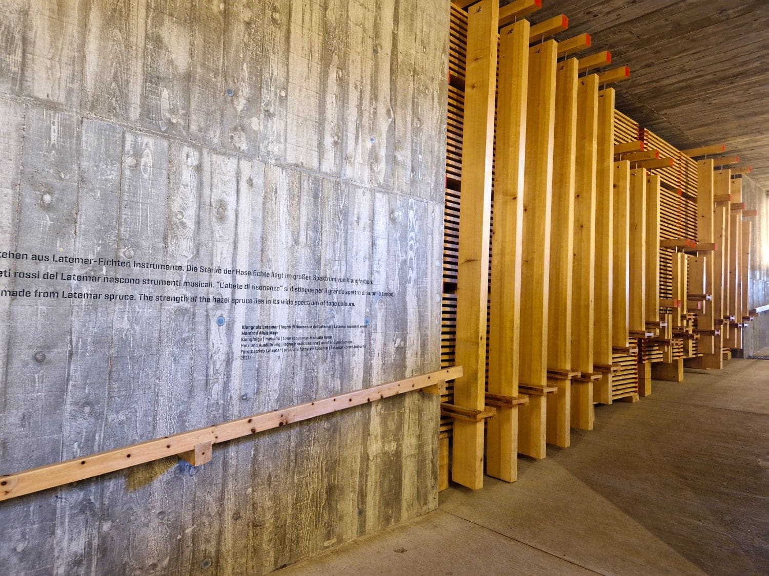 A corridor with concrete walls displaying text. The right wall is adorned with wooden beams of varying heights and colors, reminiscent of the vibrant hues found around Lake Carezza. A wooden handrail runs along the left side, adding a natural touch to the modern architectural design.