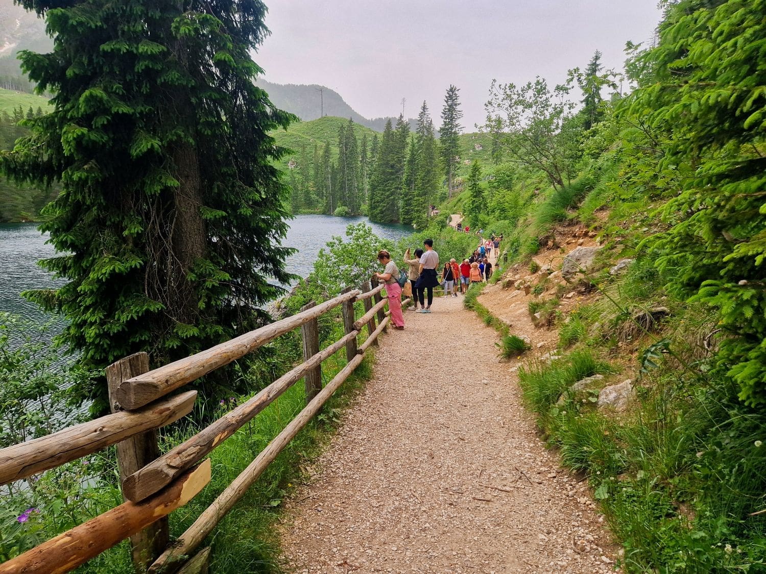 A group of people walking along a dirt path beside a scenic lake, surrounded by lush, green trees and hills. A wooden fence runs parallel to the path on the left side, overlooking the lake. The sky is slightly overcast, and the area appears serene and natural.