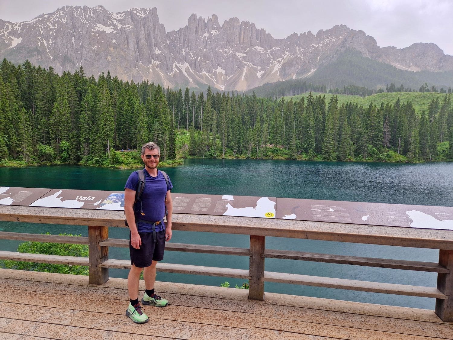 A man in a blue shirt and black shorts stands on a wooden platform overlooking a clear lake surrounded by dense green forest. Majestic, snow-capped mountains tower in the background beneath a cloudy sky. Interpretive signs are visible on the railing.