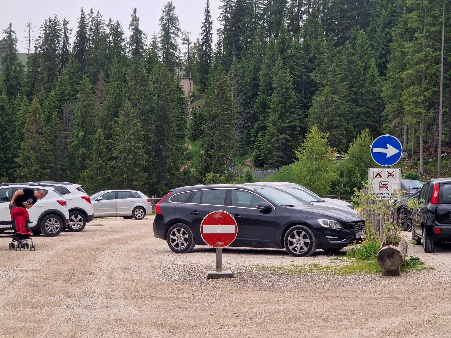 A gravel parking lot surrounded by trees. Several cars are parked, and a person is pushing a red stroller near a white SUV. A red and white no-entry sign is in the foreground, and a blue and white one-way sign is in the background.