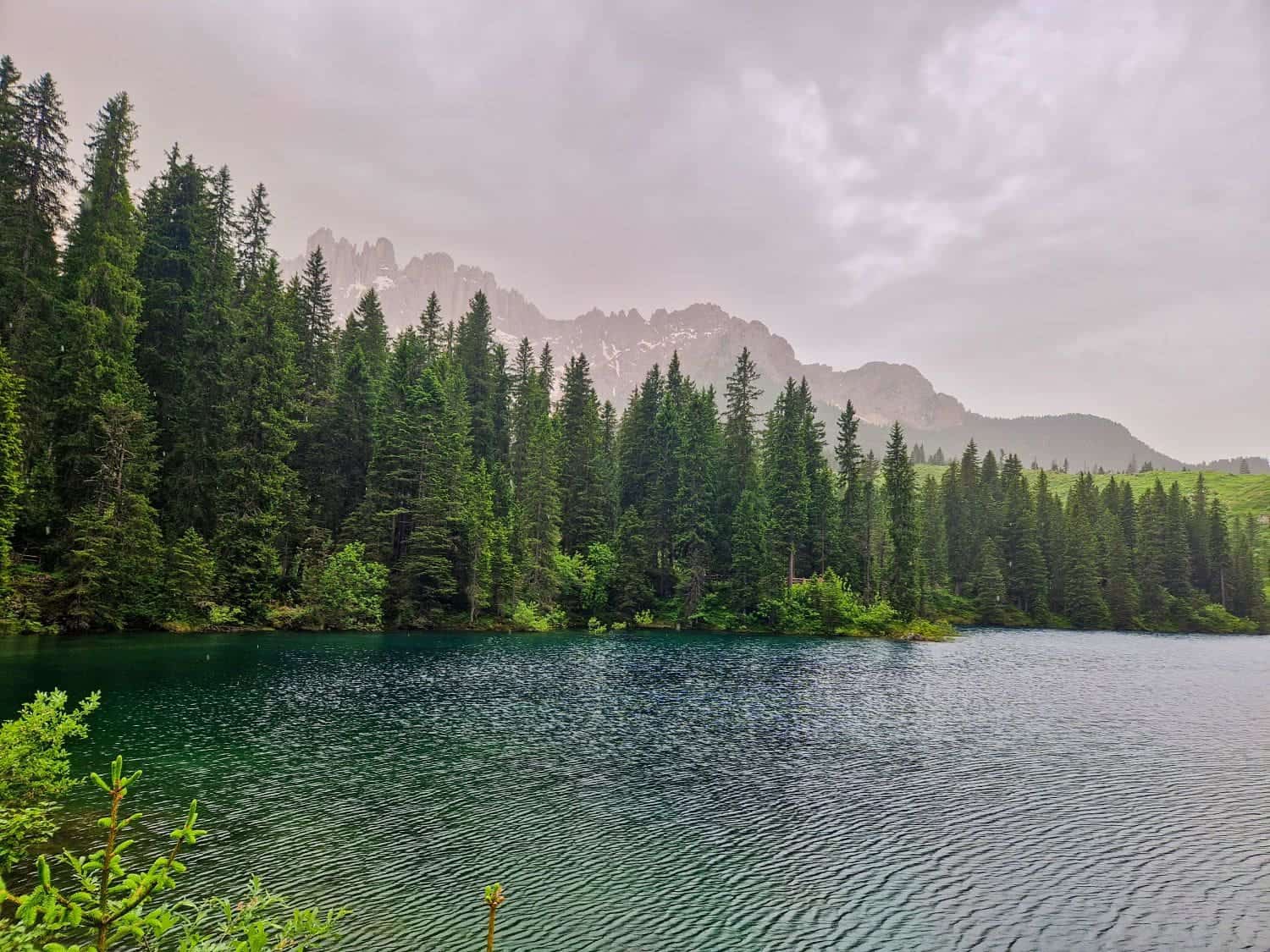 A serene mountain lake, known as Lake Carezza, is surrounded by dense evergreen trees. The water in the lake is clear with a greenish-blue hue. In the background, rugged mountain peaks are partially obscured by a cloudy and overcast sky.