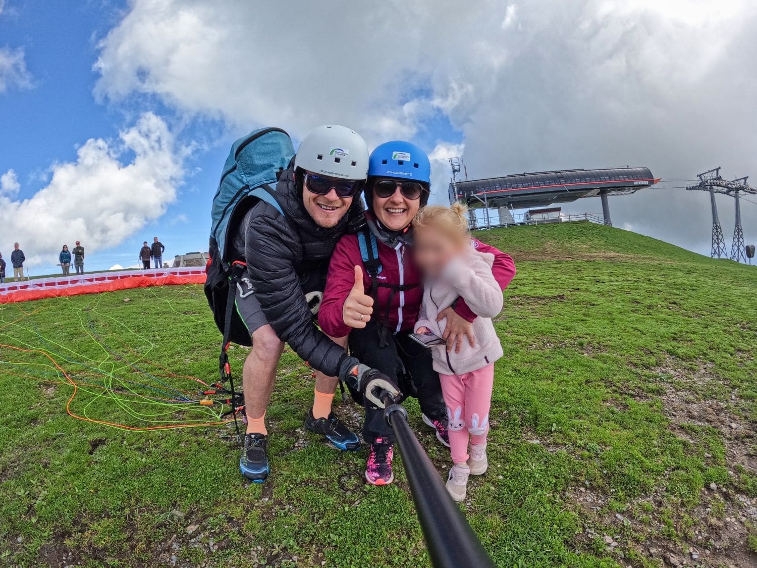 Three people on a grassy hilltop in the Dolomites, preparing for a paragliding adventure. The adults are wearing helmets and harnesses, smiling at the camera. The child, whose face is blurred, is holding onto one adult. A ski lift structure and a few people are in the background.