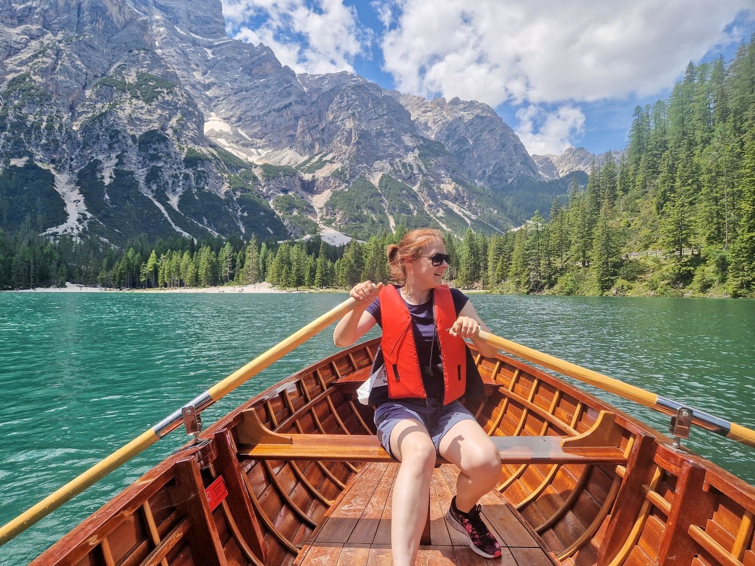 A person wearing a red life jacket and sunglasses is rowing a wooden boat on a clear turquoise Braies lake in the Dolomites surrounded by forested mountains. The sky is partly cloudy, and there are rocky mountain peaks in the background.