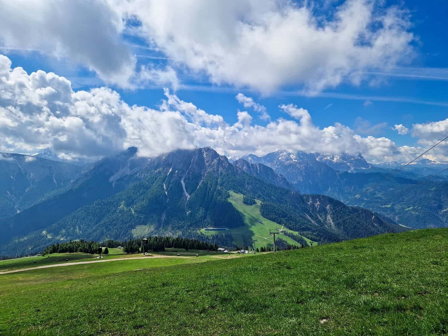 A vibrant landscape showcasing green fields in the foreground and majestic mountains in the background beneath a partly cloudy sky. The mountains are partially covered in trees and have patches of snow, with a ski lift visible on one of the slopes—an ideal spot for tandem paragliding in the Dolomites.