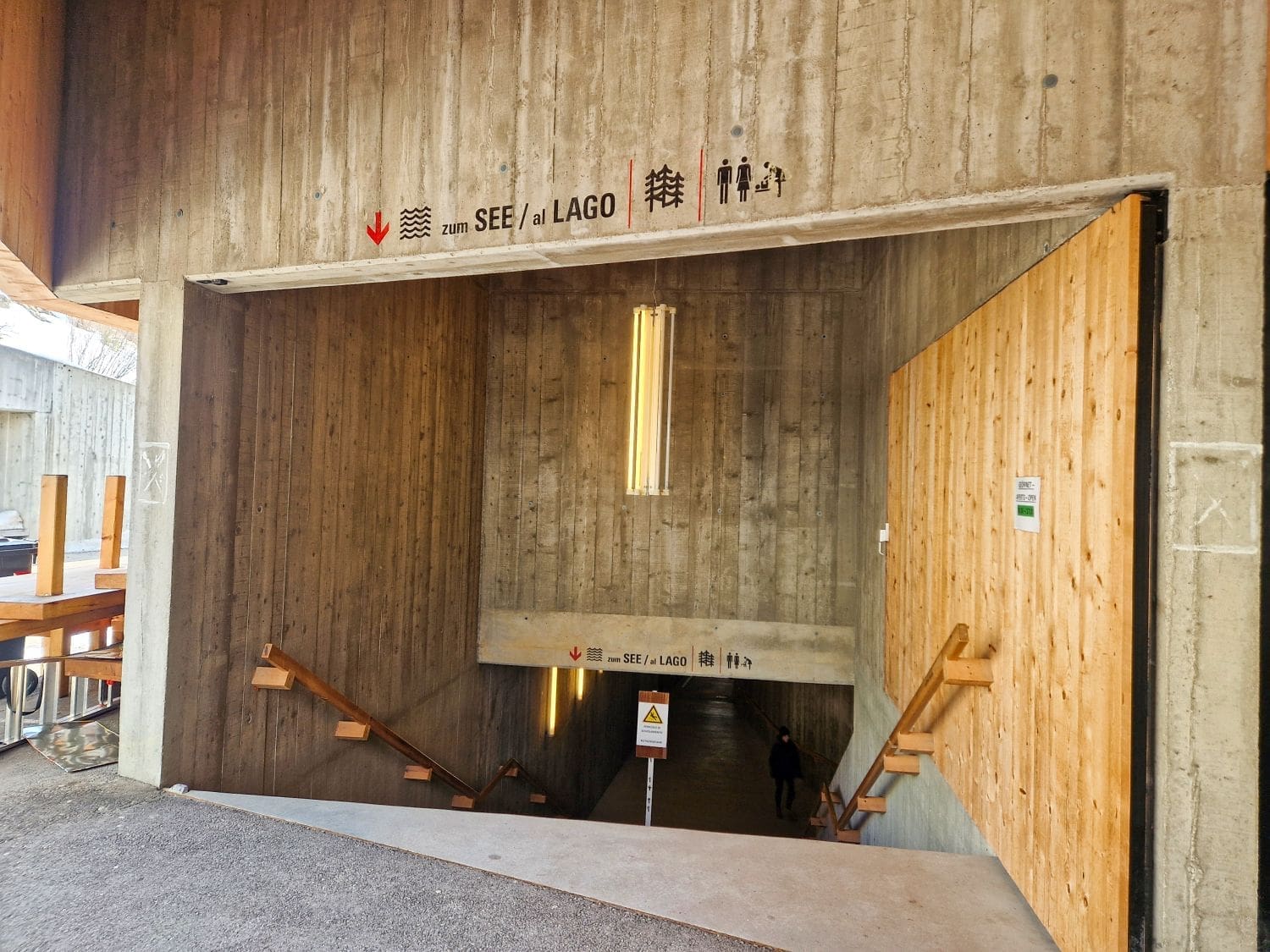 A concrete and wooden stairway leading down to an underground area marked with signs reading "SEE / al LAGO" and depictions of a restroom. Illuminated by vertical fluorescent lights, the structure features modern architecture with a clean, minimalist design reminiscent of the beauty surrounding Lake Carezza.