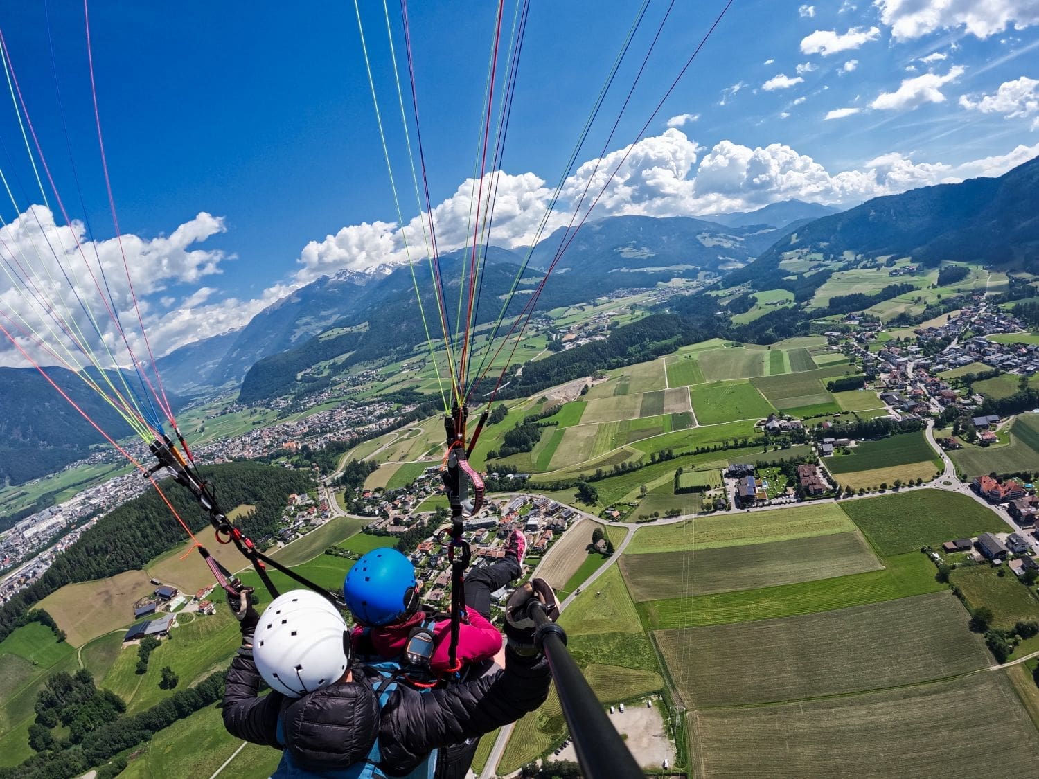 Two paragliders with colorful canopies soar high above a picturesque landscape featuring fields, villages, and mountains under a bright blue sky with scattered clouds. The view from above captures the vast expanse of green and the winding roads below.